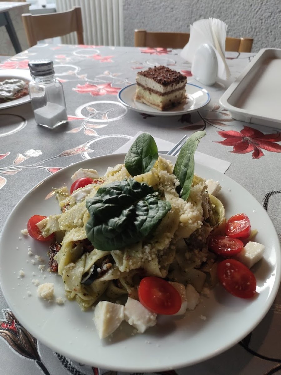 Talerz makaronu tagliatelle z pomidorkami, bazylią i serem, w tle kawałek ciasta